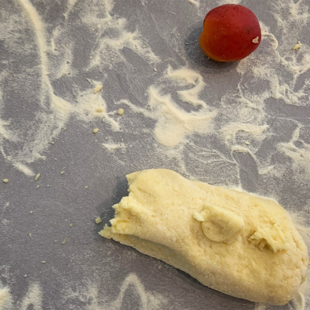 A flour-dusted kitchen counter. On it is a single apricot which is a rich orange colour. And there is a torn pile of dough.