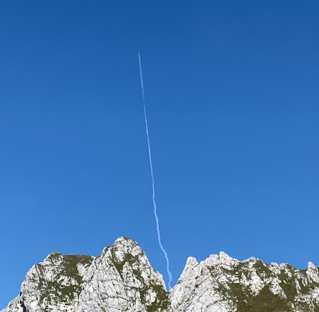 A bright blue sky and a spikey granite and-green covered mountain peak at the bottom of the image. A line of jet engine cloud runs from behind the mountain vertically to the top of the image, as if a plane is shooting from the top of the mountain.
